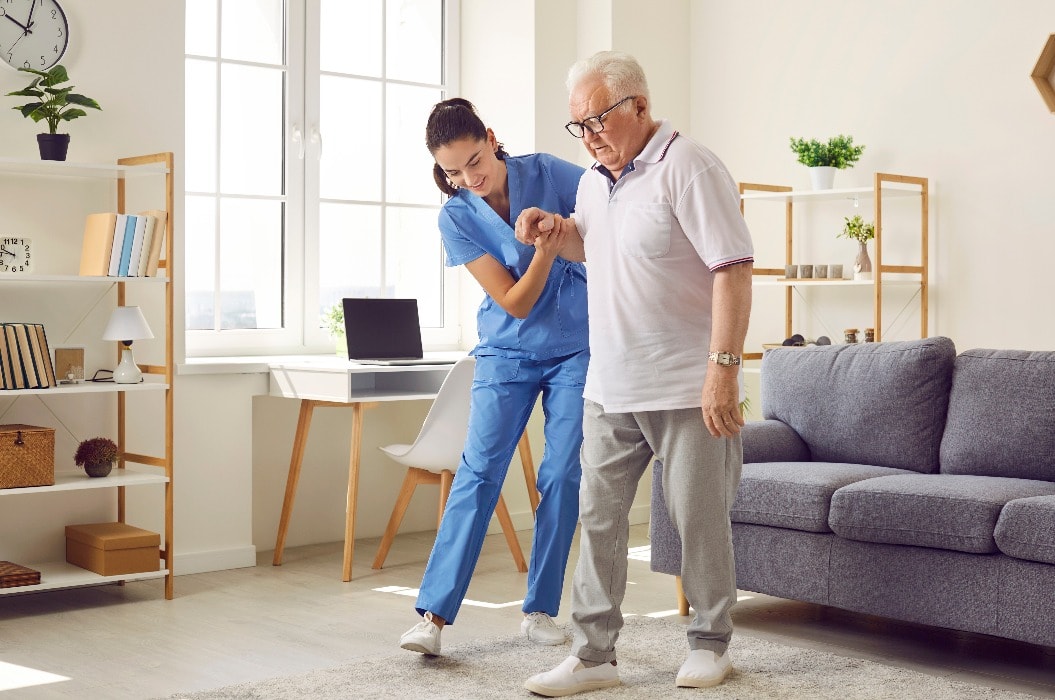 Young nurse helping elderly man walk in the room, holding his hand, supporting him. Treatment and rehabilitation after injury or stroke, life in assisted living facility, senior care concept